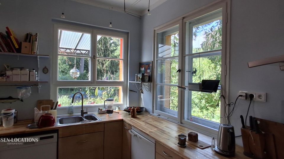 Helle Wohnküche mit hellblauen Wänden, Holzarbeitsplatten und Doppelspüle unter großen Sprossenfenstern. Offene Wandregale mit Gläsern und Vorratsdosen sowie Küchengeräte auf der Arbeitsfläche; durch die Fenster Blick ins grüne Gartenumfeld.