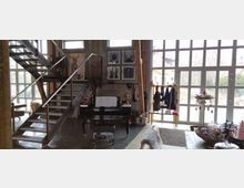 Ein modernes Wohnzimmer mit offenem Treppenaufgang aus Metall, hohen Fenstern mit Sprossen, einem schwarzen Klavier mit aufgeschlagenen Notenblättern und dekorativen Elementen. An einer Wand hängen gerahmte Kunstwerke, und durch die Fenster ist ein Außenbereich mit Gebäuden zu sehen.