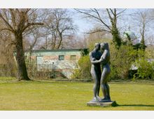 Skulptur eines sich umarmenden Paares in einem Park, umgeben von Rasen, kahlen Bäumen und Büschen; im Hintergrund ein flaches Gebäude mit Graffiti und die Kuppel eines Gebäudes.