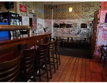 Innenraum einer kleinen Bar mit langer Holztheke und hohen Barhockern auf Holzdielenboden. Die Wände sind dicht mit Graffiti und Kritzeleien bedeckt, daneben stehen schmale Tische mit Stühlen und eine verspiegelte Wand mit Lampen.