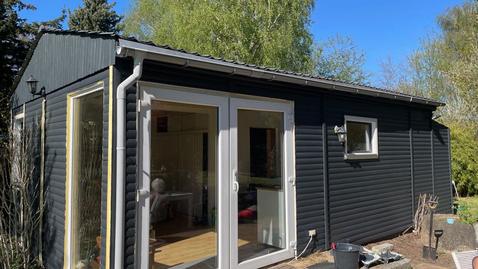 Kleines, dunkel verkleidetes Holzhaus im Garten mit flachem, leicht geneigtem Dach, Dachrinne und großen Glas-Doppeltüren sowie Seitenfenster. Vor dem Eingang liegt eine gepflasterte Terrasse; im Hintergrund stehen hohe Bäume unter blauem Himmel.