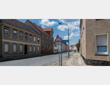 Eine ruhige Straße in einem Wohngebiet mit mehrstöckigen Häusern aus verputztem Mauerwerk und geneigten, ziegelgedeckten Dächern. Im Vordergrund ein Straßenschild mit Warnung vor Straßenschäden und der Bezeichnung „Promenade“, daneben ein Gehweg mit teils unregelmäßiger Pflasterung.