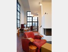 Wohnzimmer mit modernem Design, das durch hohe Decken und große Fenster mit schwarzen Rahmen geprägt ist. Im Vordergrund steht ein roter Ledersessel mit Holzarmlehnen, daneben ein weißer Kamin, im Hintergrund ein Esstisch mit Stühlen sowie eine offene Küche.