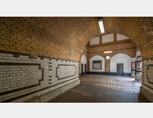 Historischer Tunnelgang mit gewölbter Decke aus sandfarbenen Ziegelsteinen und Wänden, die teils mit dekorativen weißen Kacheln versehen sind. Der Boden ist mit roten und weißen Fliesen gestaltet, und an einer Wand hängt eine analoge Uhr über einem leeren Schaukasten.