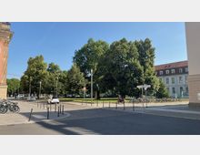 Straßenansicht mit einem kleinen Park im Hintergrund, gesäumt von großen Bäumen und einer Statue im Zentrum. Auf der rechten Seite steht ein klassisches Gebäude mit roten Dachziegeln und weißen Fassaden, während Fahrräder entlang eines Geländers geparkt sind.