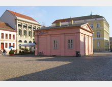 Ein gepflasterter Platz mit einem rosa Gebäude im Vordergrund, beschriftet mit „WAAGE“. Im Hintergrund stehen weitere historische Gebäude mit gelber und weißer Fassade sowie klassizistischen und barocken architektonischen Elementen.
