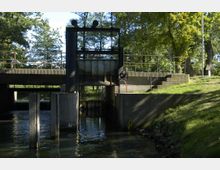 Ein kleines Schleusenbauwerk aus Beton mit Metalltoren, umgeben von Bäumen und Grasflächen. Das Wasser fließt unter der Schleuse hindurch; Treppen und ein Geländer führen zu einer Plattform oberhalb der Konstruktion.