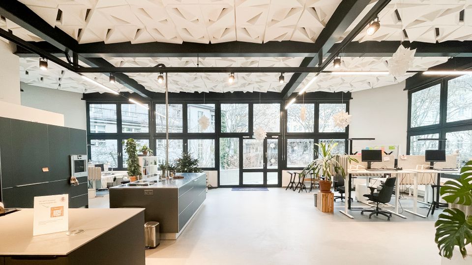 Heller, offener Büroraum mit großen Glasfenstern und Glastüren zur Außenseite, weißen Wänden und einem hellen Boden. Auffällig sind die geometrisch strukturierte Decke mit schwarzen Trägern, eine zentrale Kücheninsel sowie mehrere Arbeitsplätze mit Monitoren und Zimmerpflanzen.