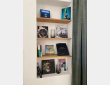 Ein in einer Wand eingebautes Regal mit drei Holzablagen, auf denen Bücher, Magazine und Dekorationsobjekte wie Glasvasen arrangiert sind. Rechts des Regals hängt ein bodenlanger, grüner Vorhang.