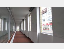 Überdachter Gang mit roten Fliesen und weißen Wänden, links eine durchgehende Fensterfront, rechts eine Reihe von großen, offenen Bögen mit Blick auf die Straße und benachbarte Gebäude. Am Ende des Gangs ist eine Glastür mit Gitter zu sehen.