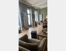 Ein Wohnzimmer mit hohen Decken, pastellgrünen Wänden und großen Fenstern mit weißen Heizkörpern dazwischen. Der Raum ist mit Holzdielenboden, gemütlichen gemusterten Sofas, einem Holztisch und dekorativen Elementen wie Pflanzen, Wandbildern und Holzobjekten eingerichtet.