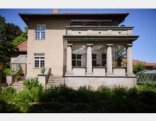 Zweistöckiges Wohnhaus mit Putzfassade und rotem Ziegeldach, davor eine erhöhte Veranda mit vier Säulen, großer Balkonbrüstung und breiten Treppenstufen. Im Vordergrund liegt ein dicht bepflanzter Garten; seitlich sind ein kleines Gewächshaus und weitere Gartenbereiche zu sehen.