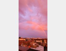 Eine Dachterrasse mit Sitzmöbeln und Pflanzen in Töpfen, im Hintergrund eine Stadtansicht mit Gebäuden unter einem rosa Himmel, über dem ein Regenbogen zu sehen ist.