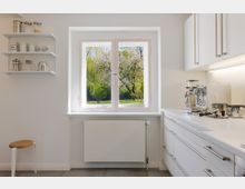 Helle, minimalistische Küche mit weißen Schränken, grauem Fliesenboden und einer langen Arbeitsplatte mit Kaffeemaschine. Ein großes Fenster über dem Heizkörper blickt auf einen grünen Garten; links sind offene Wandregale mit Gläsern und Dosen.