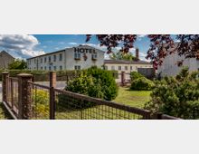 Ein schlichtes zweigeschossiges Hotelgebäude mit weißer Fassade und großen Buchstaben "HOTEL" auf der Frontseite. Im Vordergrund befinden sich ein Metallzaun, Sträucher und eine gepflegte Grünfläche, umgeben von einer ruhigen ländlichen Umgebung.