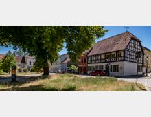 Historischer Marktplatz mit Fachwerkhäusern, gepflasterten Straßen und einer kleinen Grünfläche im Vordergrund, umgeben von Bäumen und geparkten Autos.