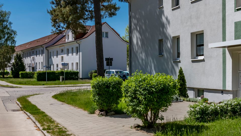 Wohnsiedlung mit mehrstöckigen, hell verputzten Wohnhäusern entlang einer gebogenen Gehweg- und Straßenführung. Vor dem rechten Gebäude sind ein Eingang mit Vordach, Grünstreifen, Hecken und einzelne Bäume zu sehen.