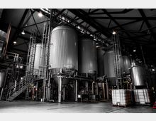 Innenansicht einer Industrieanlage mit mehreren großen, zylindrischen Tanks aus Metall, umgeben von Treppen, Rohren und Leitungen. Das Gebäude hat eine hohe, stahlverstärkte Decke mit Beleuchtung.