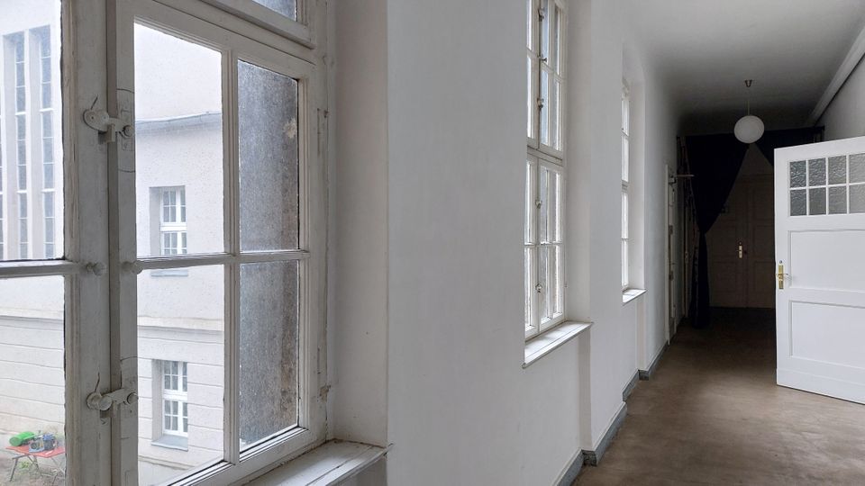 Ein heller Flur mit weißen Wänden und Holzboden, beleuchtet durch mehrere große Sprossenfenster auf der linken Seite. Rechts ist eine geöffnete weiße Tür mit Glaseinsatz zu sehen, und im Hintergrund befindet sich ein dunkler Vorhang.