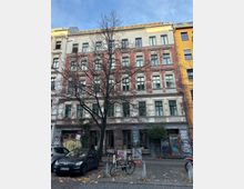 Mehrstöckiges Altbau-Wohnhaus mit rot-weiß gegliederter Fassade und vielen rechteckigen Fenstern, gesehen von der Straße aus. Im Vordergrund stehen ein kahler Baum sowie parkende Autos und Fahrräder; im Erdgeschoss sind graffitibedeckte Eingänge und Schaufenster zu sehen.