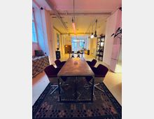 Ein moderner, loftartiger Raum mit hohen Decken, weiß gestrichenen Wänden und großen Fenstern. Im Vordergrund steht ein langer Tisch aus Metall, umgeben von gepolsterten Stühlen; im Hintergrund befinden sich Regale, ein Sofa und weitere Möbel.