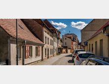 Gepflasterte Straße in einem historischen Wohnviertel mit Reihen von kleinen, verputzten Häusern mit roten Ziegeldächern und hölzernen Fensterläden. Am rechten Straßenrand sind parkende Autos zu sehen, während der Himmel klar und sonnig ist.