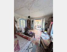 Wohnzimmer mit gemusterten Fliesenböden, rustikalen Holzmöbeln und einer niedrigen Decke; große Fenstertüren und ein Doppelfenster lassen viel Tageslicht herein. Durch die offenen Türen ist eine überdachte Terrasse mit Blick ins Grüne zu sehen.