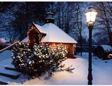 Kleines eingeschossiges Holzhäuschen mit schneebedecktem Dach, dekoriert mit leuchtenden Lichterketten; im Vordergrund verschneite Büsche und eine beleuchtete Straßenlaterne, umgeben von winterlicher Landschaft mit Bäumen.