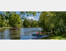 Ein ruhiger Fluss mit umgebenden grünen Bäumen, einem kleinen Steg mit abgestellten Booten im Vordergrund und im Hintergrund ein größeres Holzboot mit Passagieren. Die Szene zeigt eine idyllische Landschaft an einem sonnigen Tag.