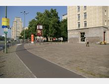 Städtische Straßenszene mit breitem Bürgersteig, separatem Radweg und modernen Gebäuden im Hintergrund. Im Vordergrund stehen eine Uhr, Werbesäulen und ein Geschäft mit der Aufschrift „Frisur u. Mode“ an einem Eckgebäude, umgeben von Bäumen.