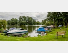Grüne Uferlandschaft an einem ruhigen Fluss mit mehreren kleinen Booten, die am Rand angelegt sind. Im Vordergrund stehen eine Holzbank und üppige Pflanzen, im Hintergrund sind Bäume und dichte Vegetation zu sehen.