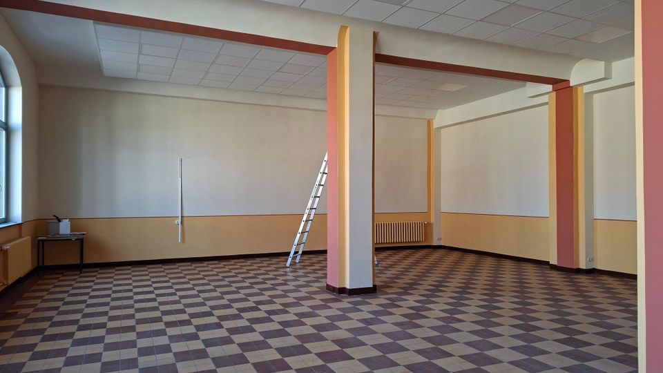 Leerer Innenraum eines größeren Raums mit kariertem Fliesenboden, abgehängter Rasterdecke und mehreren tragenden Säulen in Creme- und Rottönen. An der Wand stehen eine Leiter sowie Heizkörper und ein kleines Tischchen, links sind hohe Fenster zu sehen.