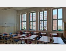Heller Klassenraum mit mehreren Reihen von Tischen und rot-blauen Stühlen auf Parkettboden, dazu Deckenleuchten und eine Garderobenleiste an der Wand. Große, hohe Fensterfront mit Fensterbänken, auf denen Topfpflanzen und Gießkannen stehen; draußen sind Wohnblocks zu sehen.