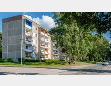 Mehrstöckiges Wohngebäude mit Balkonen, teils mit Blumen geschmückt, umgeben von Grünflächen und hohen Bäumen entlang einer gepflasterten Straße; im Hintergrund blauer Himmel mit weißen Wolken.