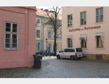 Hof in einer Altstadt mit gepflastertem Boden, umgeben von pastellfarbenen Gebäuden mit großen Fenstern; an einem Gebäude steht "Gaststätte zur Ratswaage", davor parkt ein weißer Lieferwagen und ein Müllcontainer steht an der Ecke.