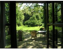 Blick von einem Fenster auf einen Garten mit grüner Wiese, Bäumen und Sträuchern. Im Vordergrund stehen mehrere Gartentische und Stühle aus Metall, teils mit Körben oder Steinobjekten dekoriert.