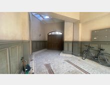 Eingangsbereich eines Altbaus mit gepflastertem Boden, einer Holztür mit Oberlicht, grünen Holzwandpaneelen und einem Fahrrad sowie Briefkästen an einer der Wände.