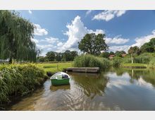 Kleiner Fluss oder Teich in einer ländlichen Umgebung mit einem grünen Ruderboot am Ufer, umgeben von Bäumen, hohen Gräsern und einem hölzernen Steg. Im Hintergrund sind Wiesen, ein Weidenbaum und Gebäude mit roten Dächern sichtbar.