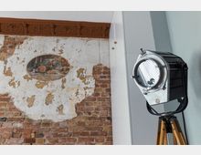 Eine detailreiche Innenraumszene mit einer freiliegenden Backsteinwand, an der ein teilweise abgenutztes Wandbild mit einem Stillleben zu sehen ist. Auf der rechten Seite steht ein großer, silberner Studiostrahler auf einem Holzstativ vor einer weißen und blauen Wand.