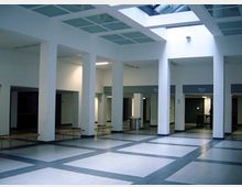 Offener Innenbereich eines modernen Gebäudes mit weißen Wänden, Säulen und einer gitterartigen Decke mit einem großen Oberlicht. Der Boden ist mit hellgrauen und dunkelgrauen Fliesen in einem quadratischen Muster gestaltet, und an den Wänden befinden sich Eingänge.