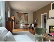 Wohn- und Essbereich mit Holzboden, großem Wandkarten-Poster und Esstisch mit vier weißen Stühlen unter hängenden Glühbirnen. Links stehen eine Kommode und ein Sofa am Fenster, rechts sind ein Durchgang zu einem weiteren Raum sowie ein Kamin und ein Sessel zu sehen.