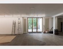 Ein leerer Raum mit Betonboden, weißen Wänden und großen Glastüren, die zu einem Außenbereich mit einer bewachsenen Steinmauer führen. Links sind Leitern an der Wand befestigt, rechts stehen Reinigungsgeräte und ein Getränkekasten.