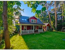 Ein rotes Einfamilienhaus im skandinavischen Stil mit weißem Balkon und Satteldach, umgeben von einem grünen Garten und Bäumen. Im Vordergrund eine Rasenfläche, im Hintergrund dichte Vegetation und Sonnenlicht.