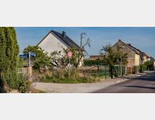 Straßenszene in einem Wohngebiet mit Einzelhäusern, teilweise umgeben von Bäumen und Zäunen. Im Vordergrund ein Verkehrsschild mit der Geschwindigkeitsbegrenzung von 30 km/h sowie ein Straßenschild an einer Weggabelung.