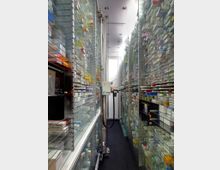 Ein automatisiertes Lagersystem mit hohen, dicht gepackten Regalen, die mit Arzneimittelpackungen gefüllt sind. In der Mitte des Ganges ist ein vertikales, motorisiertes System oder Roboterarm sichtbar, das zur Lagerung und Entnahme der Medikamente dient.