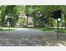 Verkehrsübungsplatz mit mehreren Fußgängerampeln, die grün leuchten, umgeben von Bäumen und einer Bank im Hintergrund; gepflasterte Wege und Begrenzungen mit Grasflächen und einem Bauzaun.