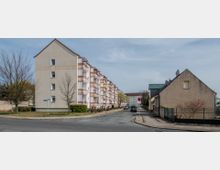 Straßenszene in einem Wohngebiet mit mehrstöckigen, weißen Wohngebäuden mit Balkonen links und einem freistehenden Einfamilienhaus mit hellbrauner Fassade rechts. Im Vordergrund führt eine asphaltierte Straße durch die Umgebung, gesäumt von vereinzelten Bäumen und geparkten Autos.