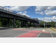 Eine Straßenkreuzung mit rot markiertem Fahrradstreifen, über der eine erhöhte Stahlbrücke für den Zugverkehr verläuft. Im Hintergrund befinden sich Bäume, ein modernes Gebäude und teils bewölkter Himmel.