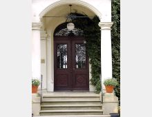 Eine Eingangstür in einem klassischen Stil mit dunklen Holztüren und verzierten Glasfenstern, eingerahmt von zwei weißen Säulen. Rechts ist eine Wand mit Efeu bewachsen, und vor der Tür stehen zwei Blumentöpfe mit Grünpflanzen auf den Stufen.