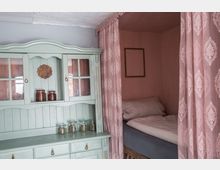 Eine kleine, gemütliche Schlafnische mit rosa Vorhängen und einem Bett, daneben ein mintgrünes Holzbuffet mit Glastüren, das mit Einmachgläsern und Dekorationen bestückt ist. Die Wand ist in einem matten Rosa gehalten.